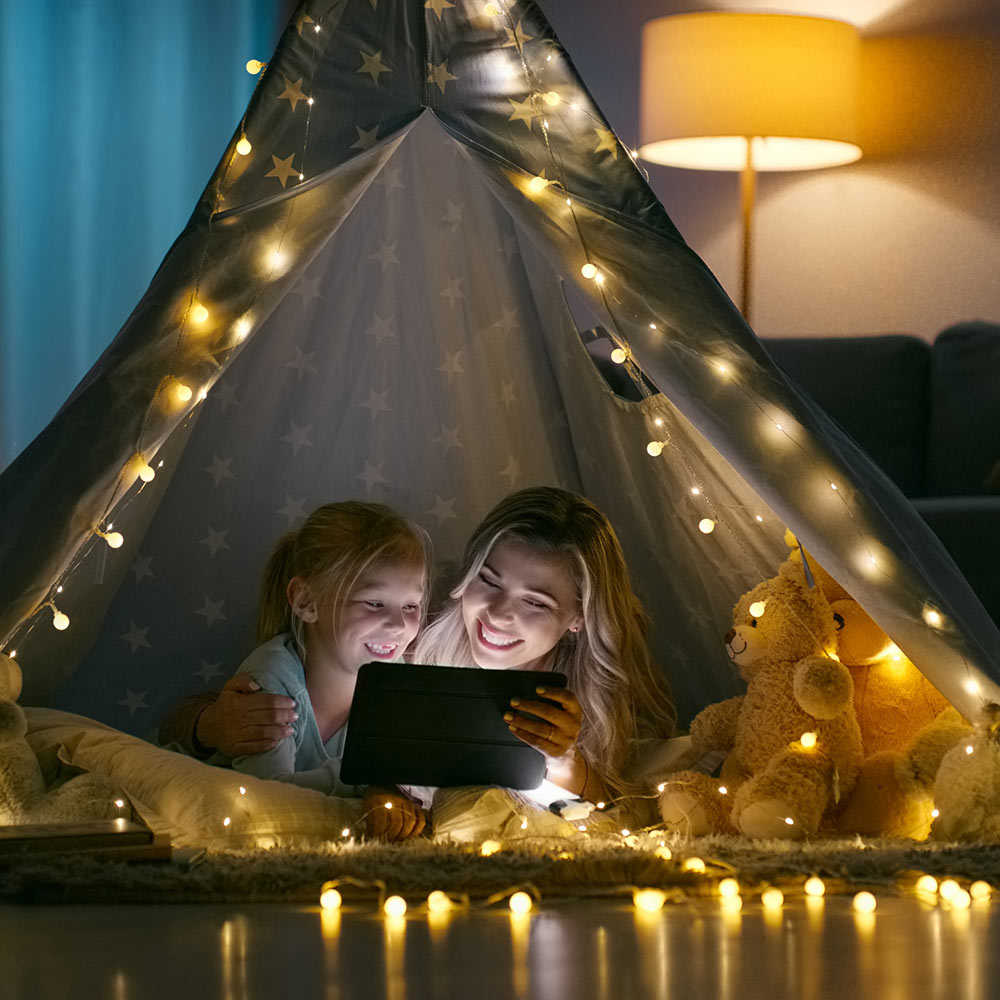 A woman and child sitting under a tent with string lights looking at a tablet