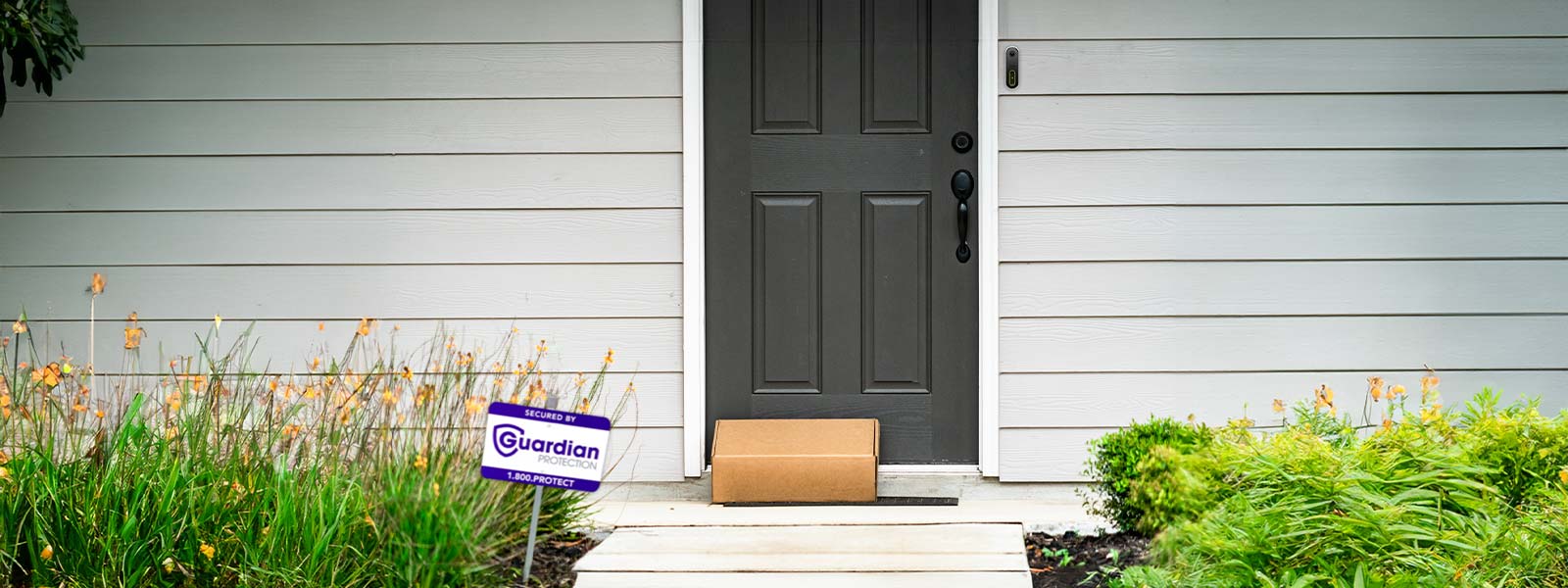 Home's front porch showing package delivered with Guardian video doorbell camera and home security yard sign